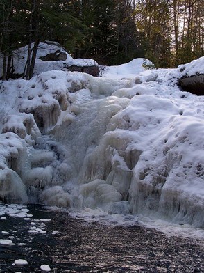 A frozen waterfall surrounded by snow-covered rocks and evergreen trees. The icy cascade appears thick and textured, with water partially flowing at the base. The scene is illuminated by soft, natural light filtering through the forest.